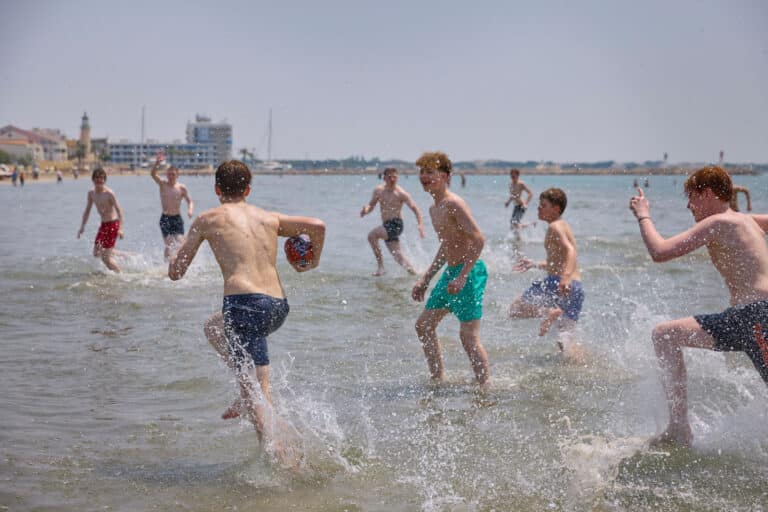 A group of boys enjoy multi-activity fun, playing with a football in shallow water at the beach, splashing and running under a clear sky. Buildings and a lighthouse are visible in the background.