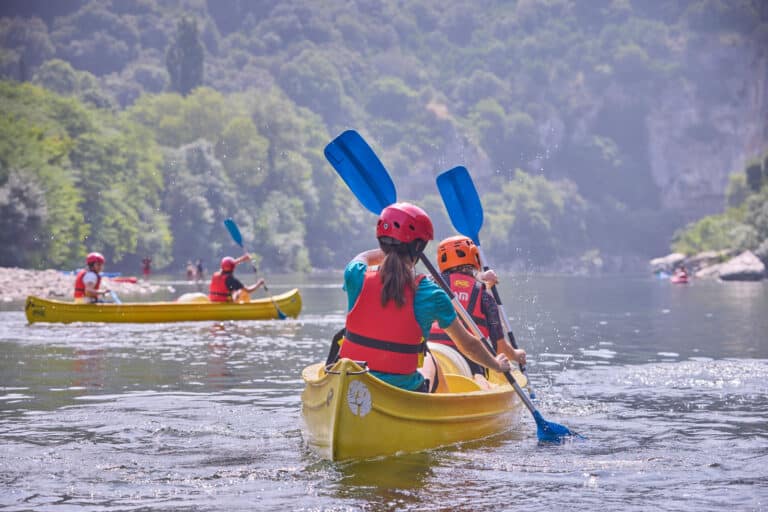 People wearing life jackets and helmets paddle yellow canoes on a calm river surrounded by green trees and hills, enjoying a multi-activity adventure in nature.