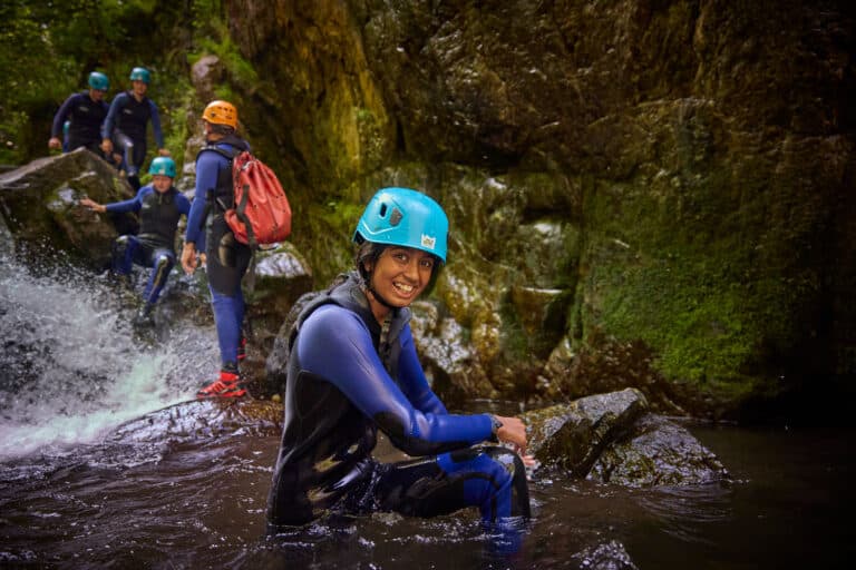 A person in a blue helmet and wetsuit sits on a rock in a stream, smiling at the camera, with three others in similar gear enjoying this multi-activity adventure near a rocky waterfall.