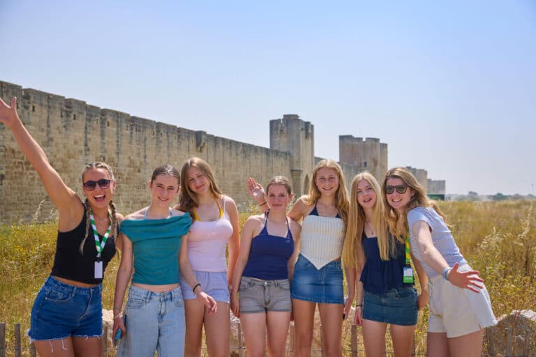 Seven young women stand smiling in front of an ancient stone wall under a clear sky, with dry grass and ruins in the background, enjoying a multi-activity day of exploration and friendship.
