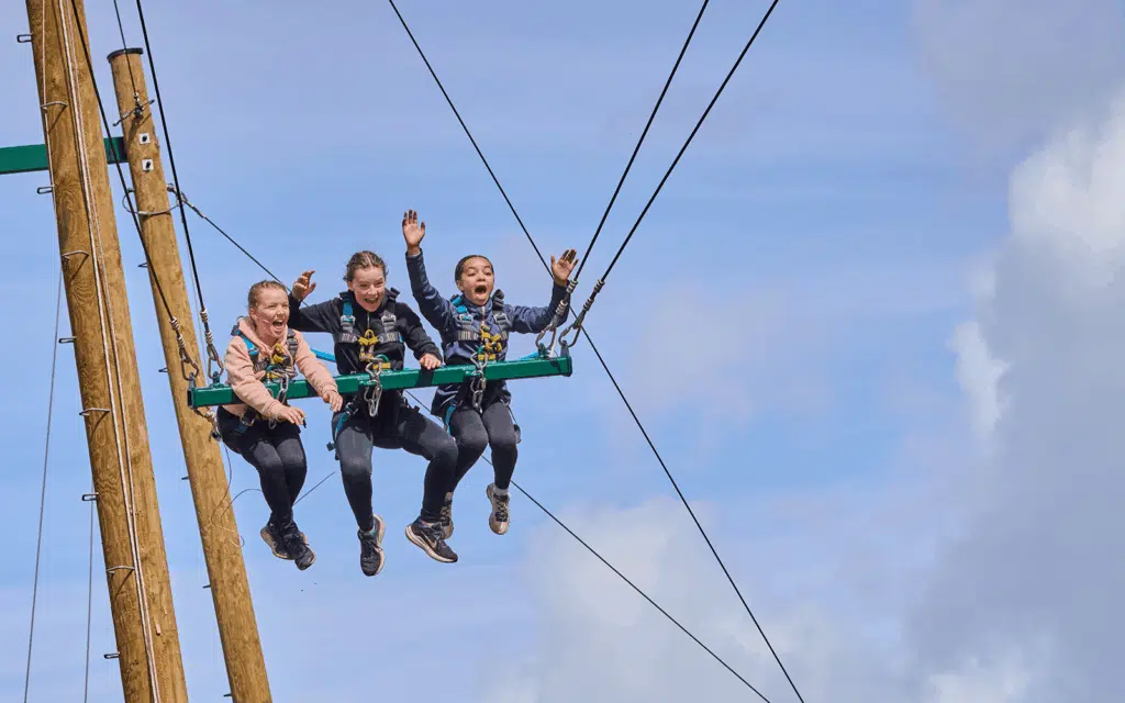 Three people wearing harnesses sit on a suspended swing high in the air, holding onto safety ropes and raising their arms against a blue sky.