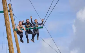 Three people wearing harnesses sit on a suspended swing high in the air, holding onto safety ropes and raising their arms against a blue sky.