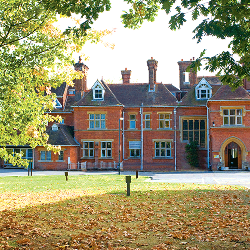 A large red-brick building with tall chimneys and multiple windows, surrounded by trees and a lawn covered with fallen leaves.