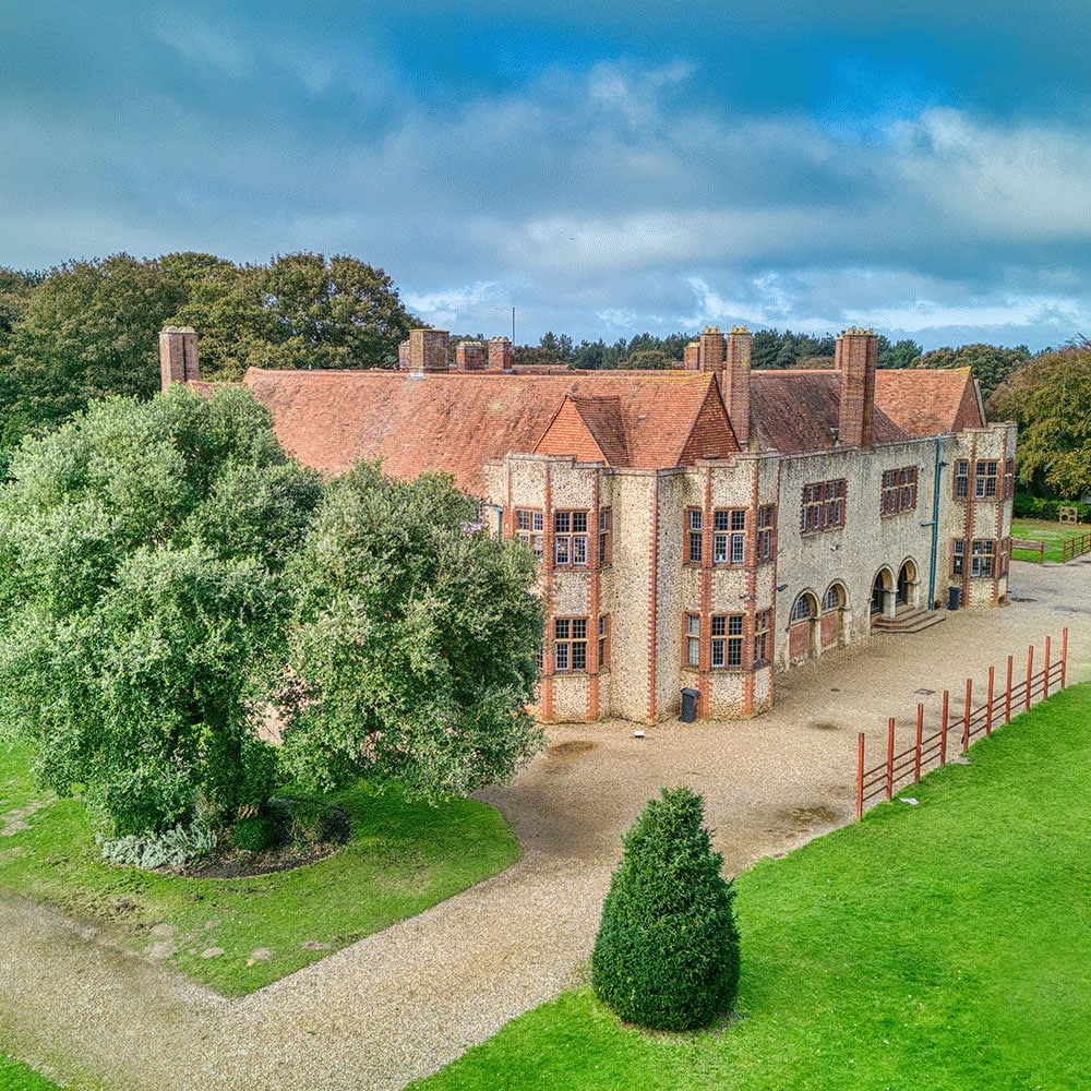 A large, historic brick building with multiple chimneys and arched windows, surrounded by green lawns, trees, and a gravel path under a partly cloudy sky.