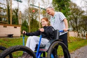 A man pushes a smiling girl seated in an adaptive wheelchair on an outdoor path, with trees and wooden buildings in the background.