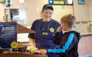 A cafeteria worker in uniform serves food to a boy at a self-service counter; the boy holds his nose while the worker smiles.