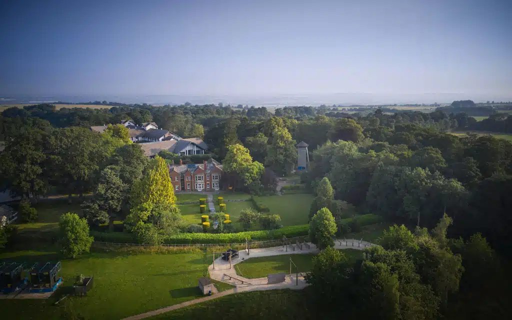 Aerial view of a large red-brick house surrounded by greenery, lawns, and trees, with a playground and paths in the foreground.
