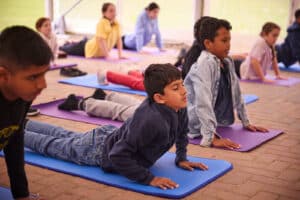 Children practice yoga poses on mats in a group setting, performing a cobra pose with hands on the floor and heads raised.