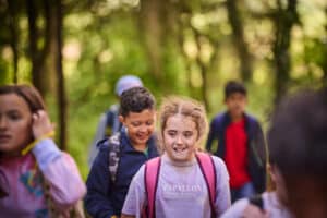 A group of children with backpacks walk together through a forested area, with trees and sunlight in the background.