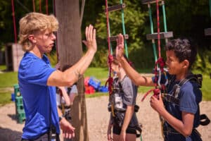 An instructor and a boy wearing safety harnesses high-five at an outdoor climbing course, with other children and climbing equipment in the background.