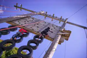 Two people climb a tire obstacle wall outdoors under a clear blue sky. They wear helmets and harnesses, participating in an adventure or ropes course activity.