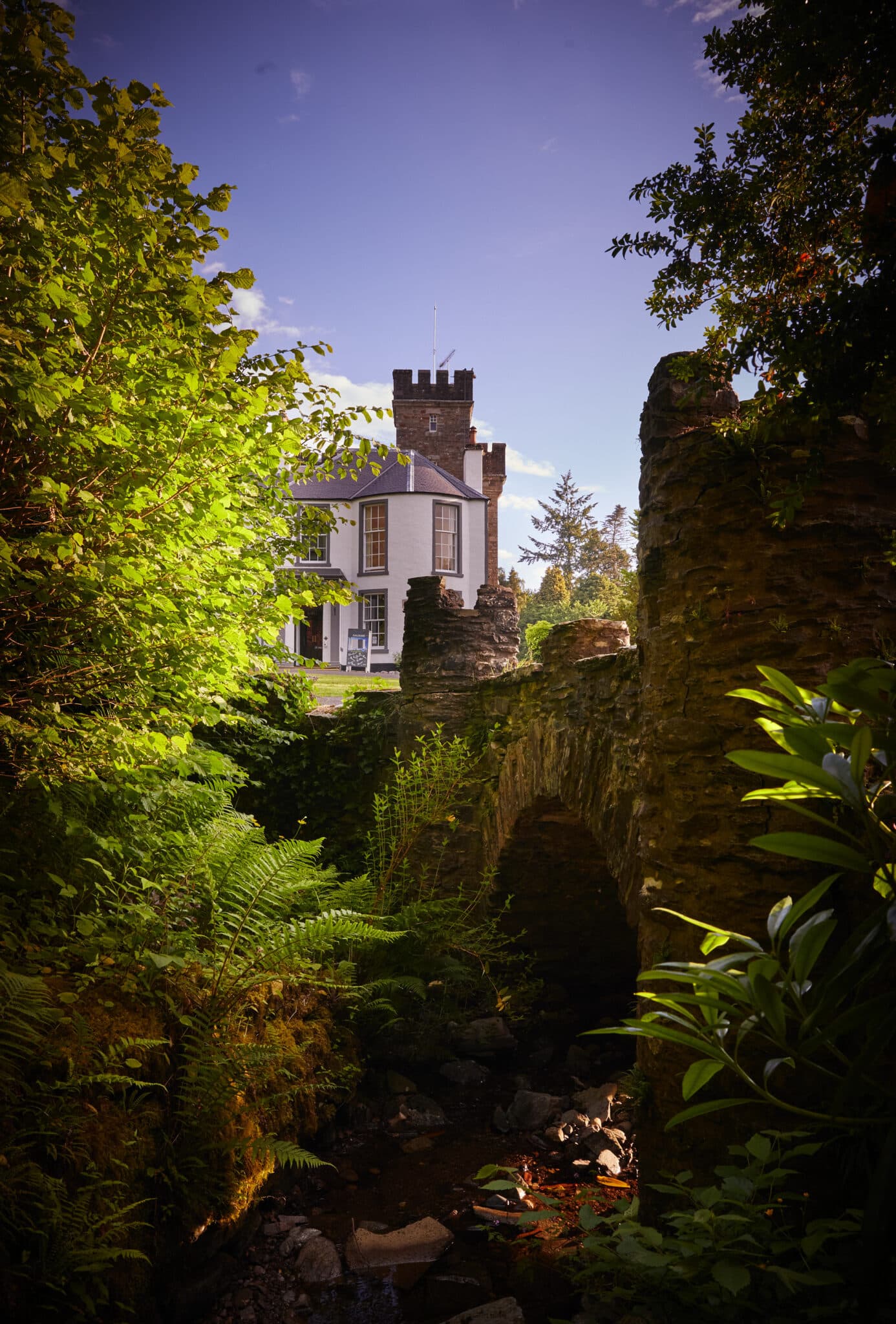 A stone bridge over a small stream is surrounded by lush greenery, with a white building featuring a tower visible in the background under a clear sky.