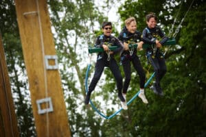 Three people in harnesses are suspended mid-air on a giant swing ride outdoors, with trees and wooden poles in the background.