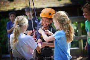 Three children adjust climbing ropes and harnesses outdoors; one child wears a helmet and smiles, while others assist, with more children blurred in the background.