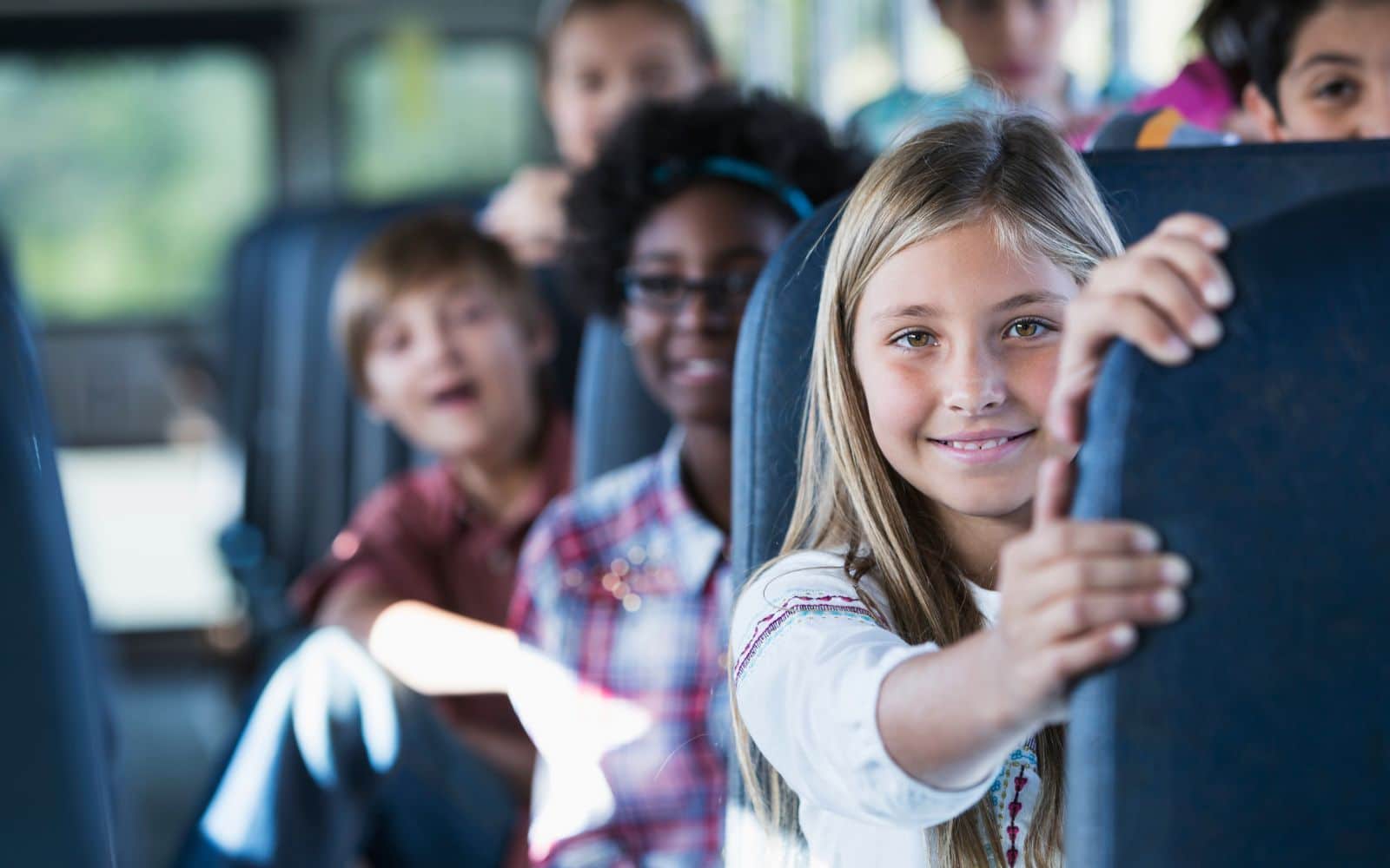 Several children are seated on a school bus. A girl in the foreground holds the seat in front of her and looks at the camera, while other children are visible in the background.