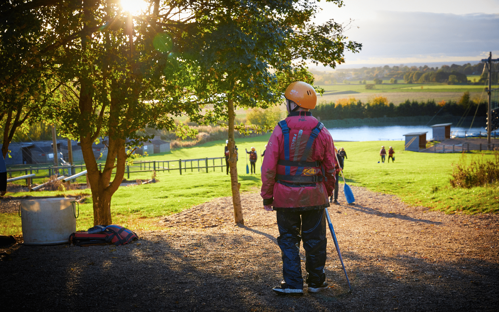 Person in outdoor gear and helmet holding paddles stands on a gravel path, looking toward a grassy area with water and other people in the distance, under a tree at sunset.