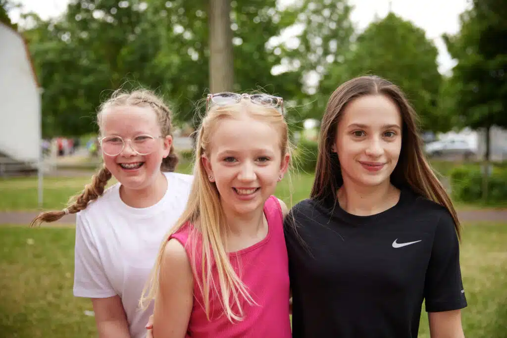 Three girls stand closely together outdoors, smiling at the camera. They are wearing casual clothing and trees are visible in the background.