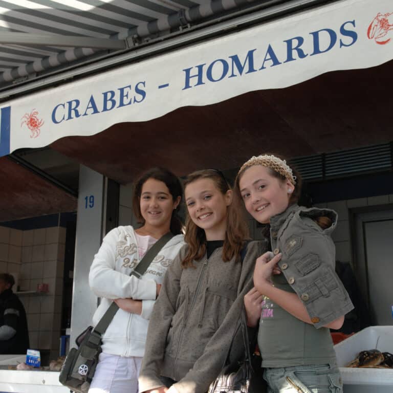 Three girls stand smiling in front of a seafood market stall with a sign that reads "CRABES - HOMARDS" under a striped awning, embracing the lively, multi-activity atmosphere around them.