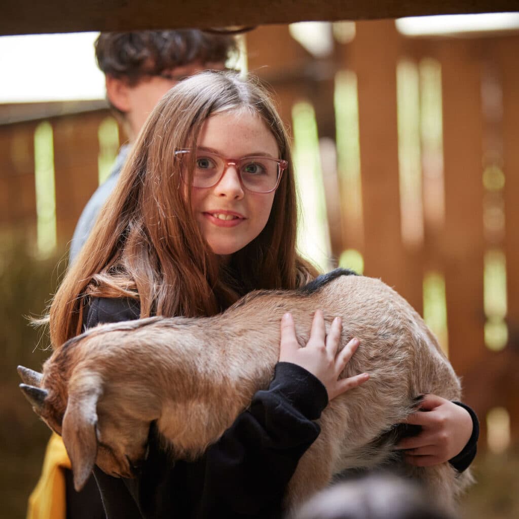 A girl with glasses holds a young goat in her arms indoors, with another person partially visible in the background.