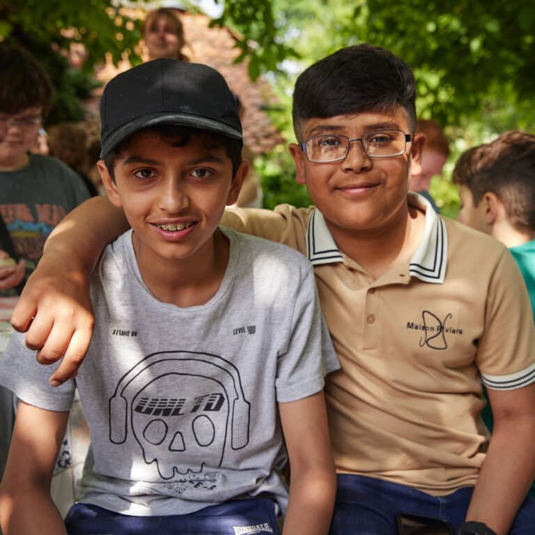 Two boys sit closely together outdoors at a multi-activity event, one with his arm around the other. They are both smiling at the camera, surrounded by other children and greenery.