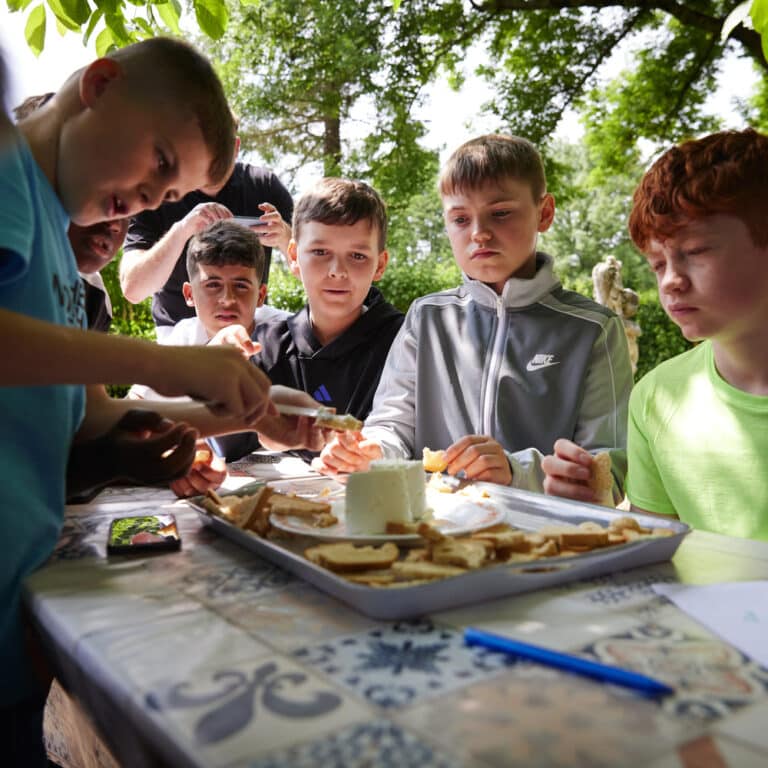 Six boys sit around an outdoor table, enjoying a multi-activity afternoon as they spread cheese or dip on bread slices from a tray. Trees and greenery are visible in the background.