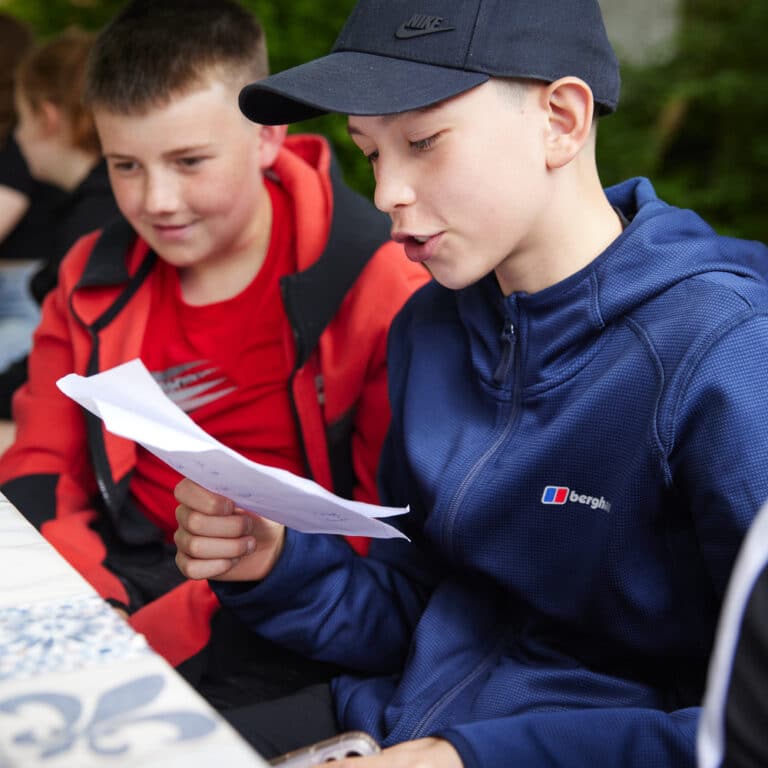 Two boys are sitting at a table outdoors, engaged in a multi-activity afternoon; one in a blue jacket and black cap reads from a sheet of paper while the other looks on.