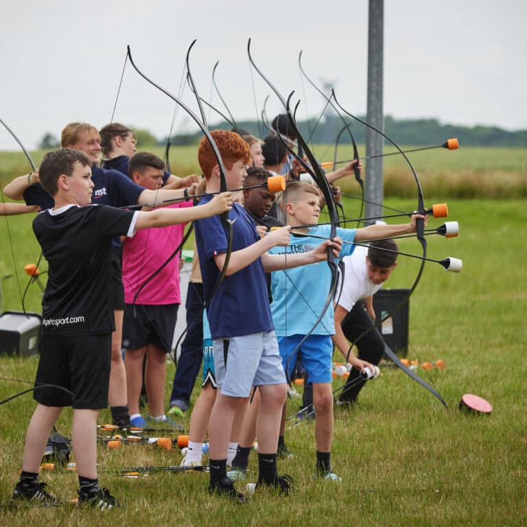A group of children stand in a row outdoors, aiming bows with foam-tipped arrows at targets during a multi-activity archery session on a grassy field.