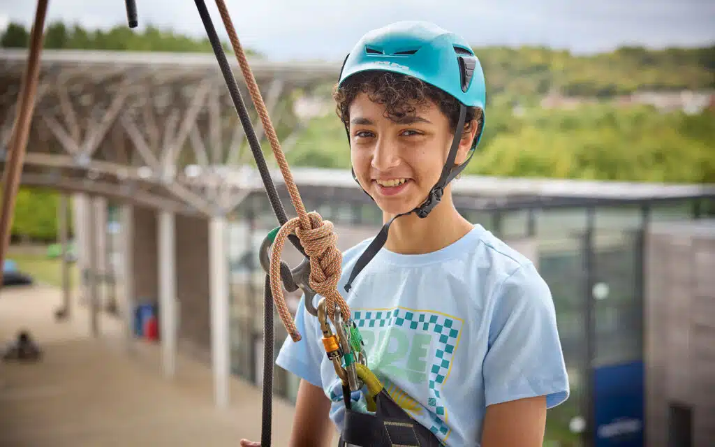 A boy wearing a blue helmet and harness stands outdoors, secured with climbing ropes, smiling at the camera during a multi-activity adventure.