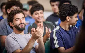 A group of young men sit closely together, some smiling and clapping, during a lively multi-activity indoor event or gathering.