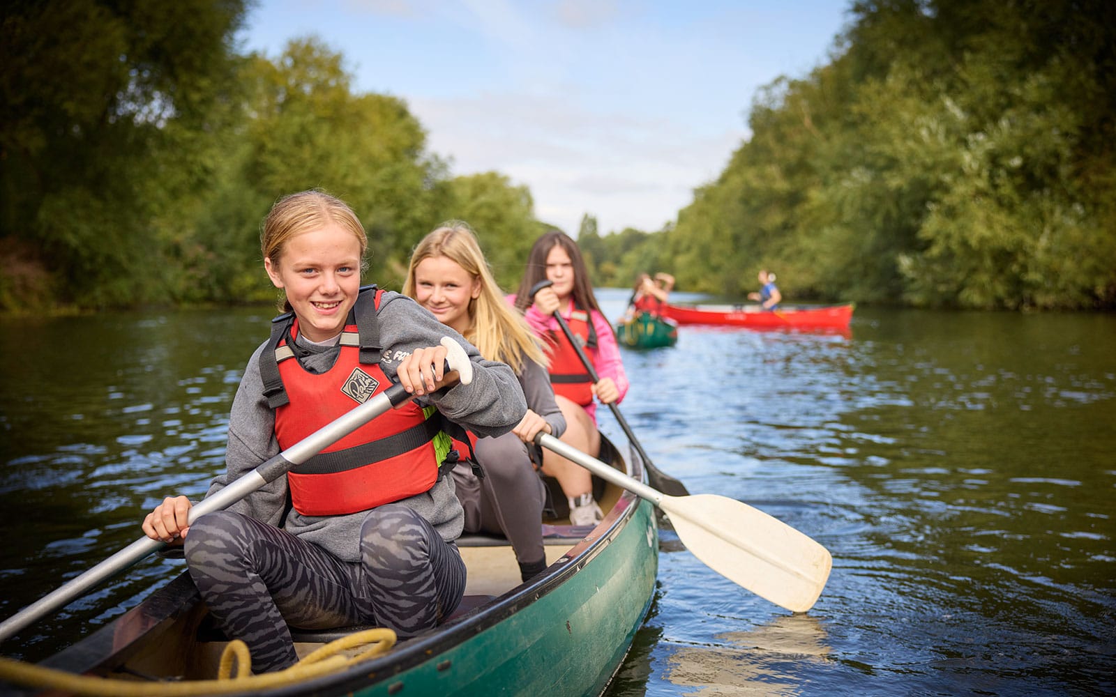 Three young people in life jackets paddle a canoe on a calm river, with another group in a red canoe visible in the background, surrounded by green trees on this exciting multi-activity adventure.