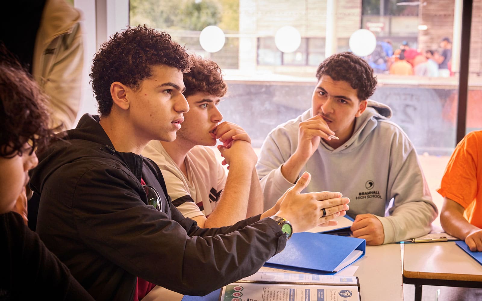 A group of students sits at a table indoors, engaged in multi-activity discussion, with one student gesturing while others listen and hold pens.