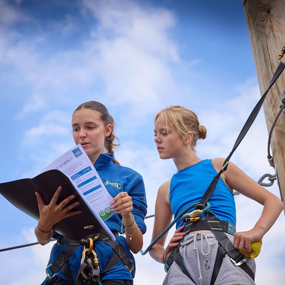 Two young people wearing harnesses stand on an outdoor ropes course, one holding a folder and papers while the other looks on, both focused on the documents during a multi-activity session.