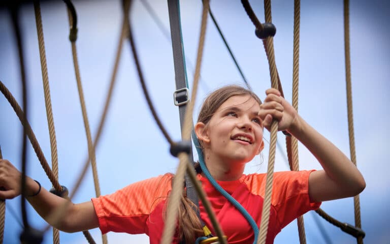A girl in an orange shirt climbs a multi-activity rope structure outdoors, looking up and smiling, with blue sky in the background.