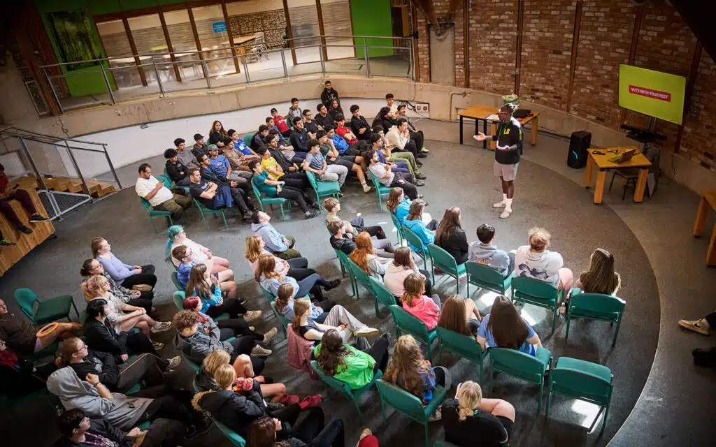 A group of people seated in rows face a speaker standing near a screen in a circular, brick-walled room with a balcony—a dynamic, multi-activity space.