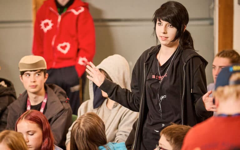 A young person in black clothing stands and speaks to a seated group of teens in a casual indoor setting, preparing them for a multi-activity session.