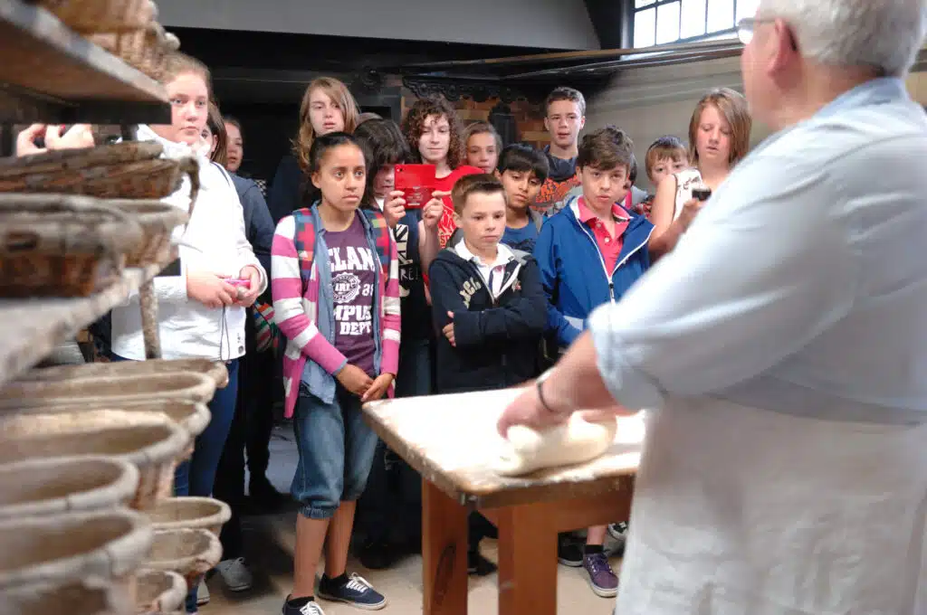 A group of children watch an older person knead dough on a table in a multi-activity bakery setting, with baskets on shelves nearby.