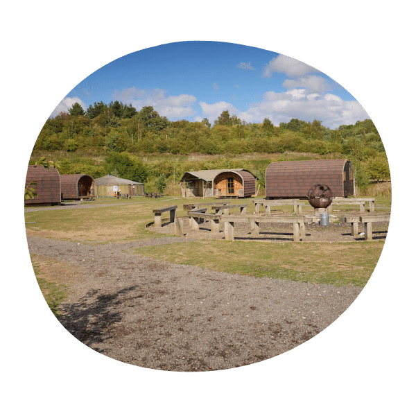 Several wooden camping pods and picnic tables are set on a grassy area with trees in the background under a partly cloudy sky.