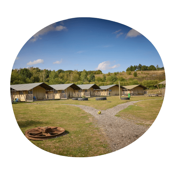 A row of canvas tents is set up on grassy land with a gravel path, surrounded by trees under a blue sky with scattered clouds.