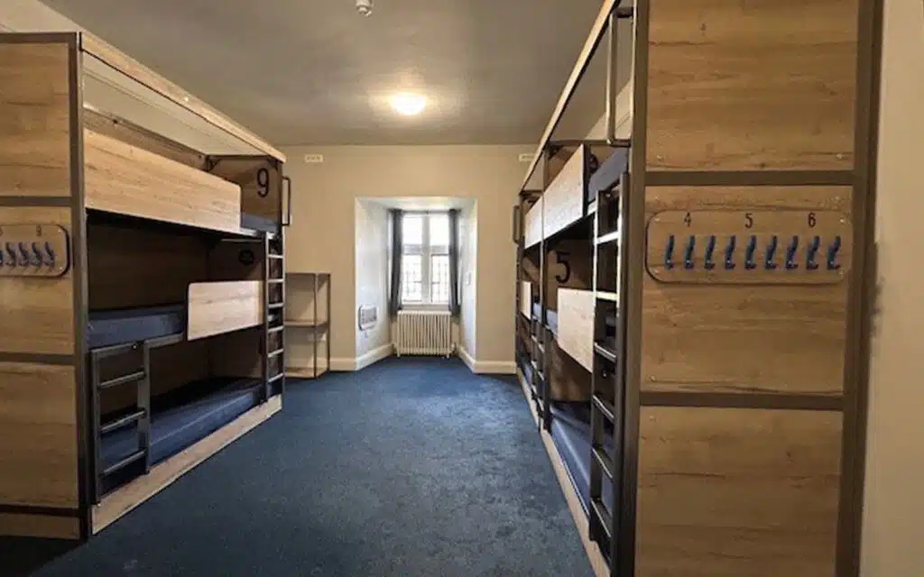 A dormitory room with wooden bunk beds, blue carpet, numbered coat hooks, and a window with natural light at the far end.