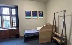 A small, sparsely furnished bedroom with a single bed, a garment rack, three posters on the wall, a window, and a trash bin.