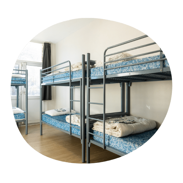 A room with metal bunk beds, blue-patterned mattresses, and folded linens, featuring a window with natural light and wooden flooring.