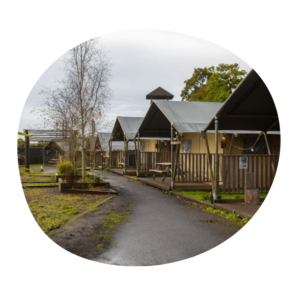 A row of canvas glamping tents with porches and benches, set along a paved path in a green outdoor area under a cloudy sky.