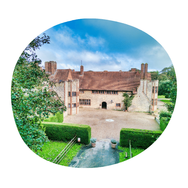 A large, historic stone manor house with a red-tiled roof, surrounded by greenery and a courtyard under a partly cloudy sky.