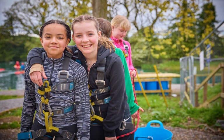 Two girls wearing harnesses stand close together and smile at the camera outdoors, with other children and safety equipment visible in the background.