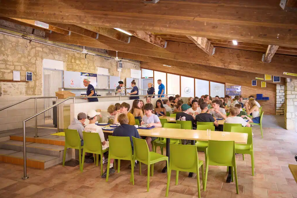 Groups of people sit at green tables eating in a cafeteria with stone walls and a wooden beam ceiling, while staff serve food at the counter.
