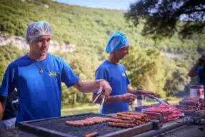 Two people in blue shirts and hairnets cook hot dogs and meat on a large outdoor grill, with trees and hills in the background.