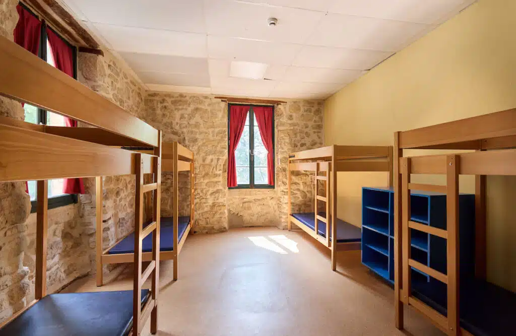 A dormitory room with stone walls, six wooden bunk beds with blue mattresses, red curtains, and shelf units, illuminated by natural light from a central window.