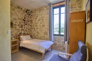 A small bedroom with a single bed, stone walls, a wooden nightstand, a window with gray curtains, a coat rack, and a gray chair.