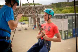 Two people in helmets and harnesses prepare for abseiling on a building exterior; one instructs while the other grasps the rope, with modern buildings and greenery in the background.