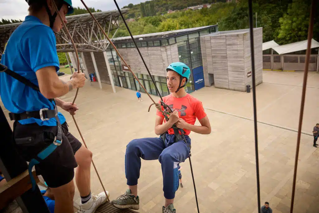 A person in a helmet and harness prepares to rappel down a building while another person assists, with a modern building and courtyard in the background.